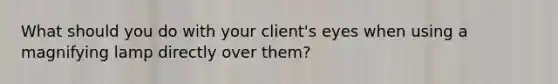 What should you do with your client's eyes when using a magnifying lamp directly over them?