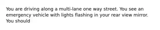 You are driving along a multi-lane one way street. You see an emergency vehicle with lights flashing in your rear view mirror. You should