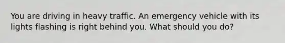 You are driving in heavy traffic. An emergency vehicle with its lights flashing is right behind you. What should you do?