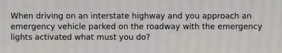 When driving on an interstate highway and you approach an emergency vehicle parked on the roadway with the emergency lights activated what must you do?