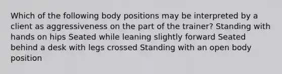 Which of the following body positions may be interpreted by a client as aggressiveness on the part of the trainer? Standing with hands on hips Seated while leaning slightly forward Seated behind a desk with legs crossed Standing with an open body position