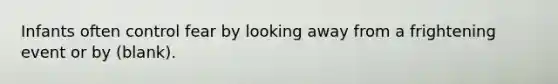 Infants often control fear by looking away from a frightening event or by (blank).