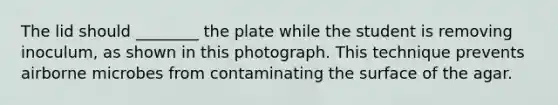 The lid should ________ the plate while the student is removing inoculum, as shown in this photograph. This technique prevents airborne microbes from contaminating the surface of the agar.