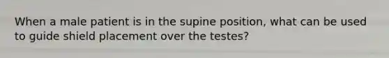 When a male patient is in the supine position, what can be used to guide shield placement over the testes?