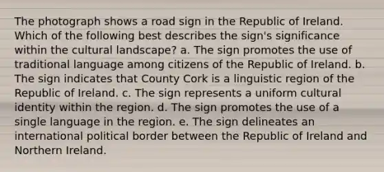 The photograph shows a road sign in the Republic of Ireland. Which of the following best describes the sign's significance within the cultural landscape? a. The sign promotes the use of traditional language among citizens of the Republic of Ireland. b. The sign indicates that County Cork is a linguistic region of the Republic of Ireland. c. The sign represents a uniform cultural identity within the region. d. The sign promotes the use of a single language in the region. e. The sign delineates an international political border between the Republic of Ireland and Northern Ireland.