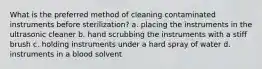 What is the preferred method of cleaning contaminated instruments before sterilization? a. placing the instruments in the ultrasonic cleaner b. hand scrubbing the instruments with a stiff brush c. holding instruments under a hard spray of water d. instruments in a blood solvent