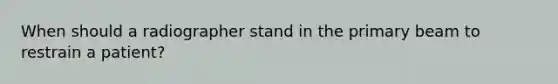 When should a radiographer stand in the primary beam to restrain a patient?