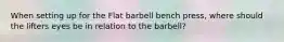 When setting up for the Flat barbell bench press, where should the lifters eyes be in relation to the barbell?