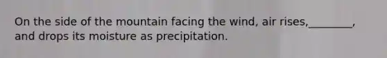 On the side of the mountain facing the wind, air rises,________, and drops its moisture as precipitation.