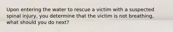 Upon entering the water to rescue a victim with a suspected spinal injury, you determine that the victim is not breathing, what should you do next?