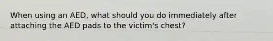 When using an AED, what should you do immediately after attaching the AED pads to the victim's chest?